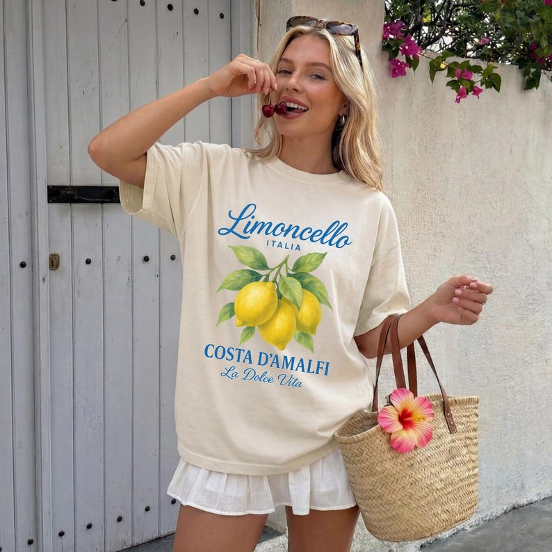 May include: Cream-colored t-shirt with a "Limoncello Italia" design featuring lemons and leaves. The shirt also has "Costa D'Amalfi La Dolce Vita" text. A straw tote bag with a pink flower is held in the hand.