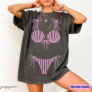 May include: A charcoal gray short-sleeve t-shirt featuring a light pink graphic print of a bikini with seashell-shaped cups and striped bottoms. The design includes thin tie-side details on the bikini top and bottom pieces.
