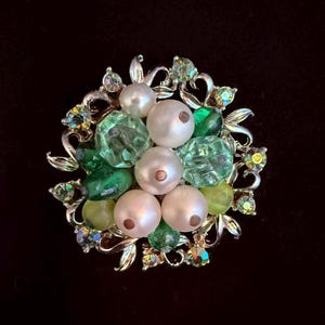May include: A vintage brooch with a floral design. The brooch features a cluster of white faux pearls, green and light green faceted glass beads, and small iridescent rhinestones set in a silver-tone metal. The brooch is set against a black background.