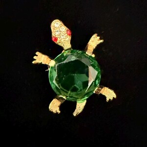 May include: A gold-toned turtle brooch with a large, faceted green crystal shell. The turtle's head and feet are detailed with gold metal and red accents. The brooch is set against a black background.