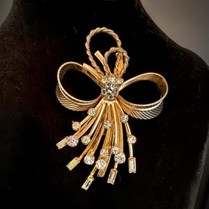 May include: A gold-toned vintage brooch with a bow design. The brooch features a central cluster of clear rhinestones and smaller stones along the radiating lines. The bow has a textured, twisted rope design and the brooch is set against a black background.