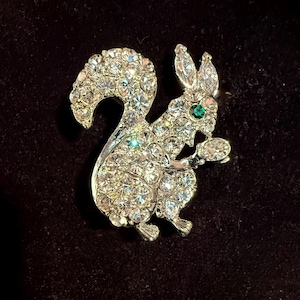 May include: A silver-toned squirrel brooch encrusted with clear rhinestones. The squirrel is depicted in a seated position, holding a small nut. The eye is a single green stone. The brooch has a classic, vintage style.
