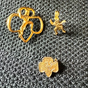May include: Three gold-colored Girl Scout pins. The largest pin features the Girl Scout logo. The other two pins are a small charm and a flower-shaped pin with the letters "GS" and an eagle emblem.