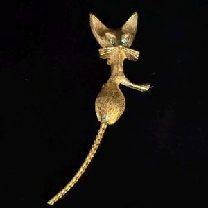 May include: A gold-toned cat brooch with a textured finish. The cat has pointed ears, a bow tie, and turquoise-colored eyes. The brooch features a long, curved tail and a slender body, creating an elegant silhouette.