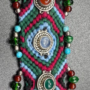 May include: Close-up of a woven bracelet with a geometric pattern in green, blue, red, and black. The bracelet features three silver-toned circular settings, each holding a different coloured stone: orange, blue, and green. Small beads in various colours and silver accents adorn the sides.