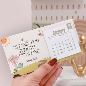 May include: A rectangular desk calendar for January 2027 featuring a light green and white colour scheme. The left side displays the text "STAND FOR THRUTH, even ALONE" and "JOSHUA 1:9" with illustrations of a sun, bee, garden sign, and watering can.