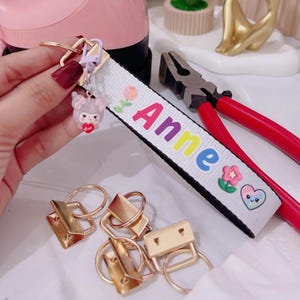 Könnte beinhalten: Ein weißes Schlüsselband mit dem Namen "Anne" in bunten Buchstaben, verziert mit Blumen und einem herzförmigen Gesicht. Der Schlüsselanhänger enthält einen goldfarbenen Verschluss und eine kleine, rosa Figur, die ein Herz hält. Auch goldfarbene Metallbeschläge sind sichtbar.