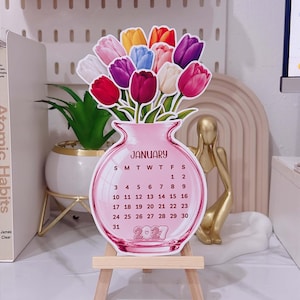 Könnte beinhalten: Ein gestanzter Tischkalender für Januar 2027 in Form einer rosa Glasvase, gefüllt mit einem Strauß bunter Tulpen in Rot-, Lila-, Weiß-, Gelb- und Blautönen. Der Kalender wird auf einer kleinen hölzernen Staffelei präsentiert.