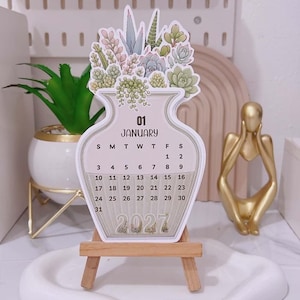 May include: A die-cut desk calendar shaped like a vase filled with various succulents. The calendar displays the month of January 2027 with a standard grid of dates. It is supported by a small wooden easel stand, featuring a neutral colour palette.