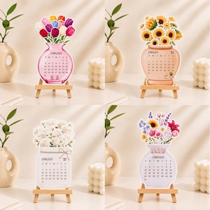 May include: Four desktop calendars displayed on small wooden easels. Each calendar features a unique floral arrangement in a vase, including tulips, sunflowers, white roses, and mixed wildflowers, with the month of January 2027 printed on each.