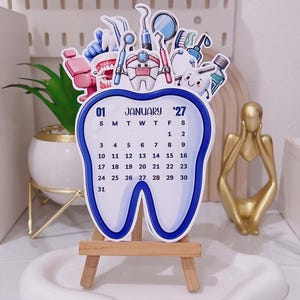 May include: A tooth-shaped desk calendar for January 2027, featuring a blue border and illustrations of dental tools, a dental chair, and a smiling tooth character. The calendar is displayed on a small wooden easel stand.