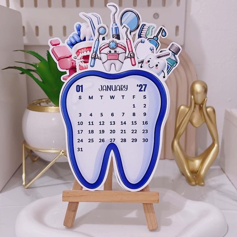 May include: A tooth-shaped desk calendar for January 2027, featuring a blue border and illustrations of dental tools, a dental chair, and a smiling tooth character. The calendar is displayed on a small wooden easel stand.