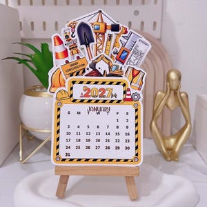 Può includere: Un calendario da scrivania per gennaio 2027 con illustrazioni a tema edilizio tra cui una gru, un escavatore, una pala, un giubbotto di sicurezza e un casco. Il calendario è esposto su un piccolo cavalletto in legno con bordo giallo.