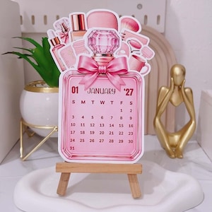 Può includere: Un calendario da scrivania fustellato a forma di flacone di profumo con un fiocco rosa, che mostra il mese di gennaio 2027. Il design presenta illustrazioni di prodotti di bellezza come pennelli e flaconi, poggiato su un piccolo cavalletto in legno.