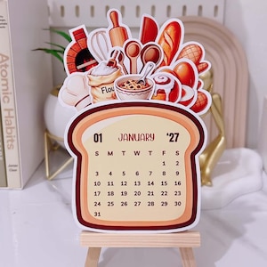 May include: A die-cut calendar shaped like a slice of bread, displaying January 2027. The top features illustrations of a brick oven, flour bag, rolling pin, whisk, wooden spoons, bread loaf, and cinnamon roll. It rests on a small wooden easel.