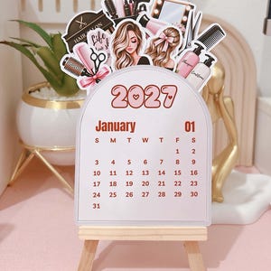 Puede incluir: Un calendario de enero de 2027 sobre un pequeño caballete de madera. La parte superior presenta ilustraciones de herramientas de peluquería, como tijeras, peine, champú, acondicionador, secador y espejo, junto a dos figuras con cabello largo.