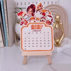 Può includere: Una scheda calendario di gennaio 2027 esposta su un piccolo cavalletto in legno. La scheda presenta uno sfondo color crema con bordi e testo arancioni. Sopra la griglia del calendario ci sono illustrazioni colorate di una persona, una volpe, funghi e una tazza di caffè.