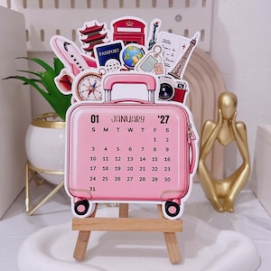 May include: A pink suitcase-shaped calendar for January 2027, displayed on a small wooden easel. The calendar features travel-themed illustrations including a passport, globe, aeroplane, camera, compass, and landmarks like the Eiffel Tower and a red phone box.