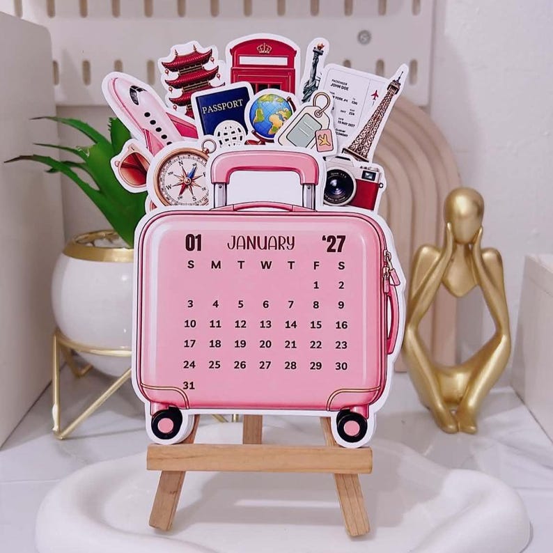 May include: A pink suitcase-shaped calendar for January 2027, displayed on a small wooden easel. The calendar features travel-themed illustrations including a passport, globe, airplane, camera, compass, and landmarks like the Eiffel Tower and a red phone booth.