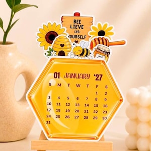 May include: A hexagonal desk calendar for January 2027 with a yellow honey-themed design. The top features a wooden sign reading BEE LIEVE in YOURSELF, a sunflower, a beehive, a bee, and a honey dipper. The calendar grid displays dates 1 through 31.