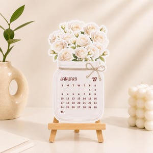 May include: A die-cut desk calendar shaped like a mason jar filled with cream-colored roses. The calendar displays the month of January 2027 with dark brown text and numbers. It is supported by a small, light-colored wooden easel stand.