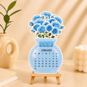 May include: A die-cut 2027 January calendar shaped like a light blue vase filled with blue roses. The calendar is displayed on a small wooden easel. The text includes the year 2027, the month January, and the days of the month from 1 to 31.
