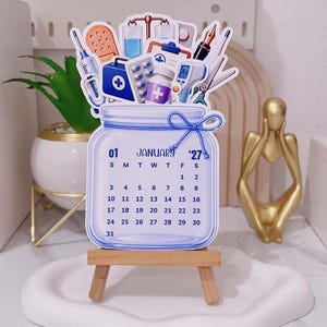 Può includere: Un calendario fustellato per gennaio 2027 a forma di barattolo di vetro con un fiocco di spago blu. La parte superiore presenta illustrazioni di forniture mediche tra cui una siringa, una benda, una sacca per flebo, un flacone di pillole, un termometro e forbici. Poggia su un cavalletto in legno.
