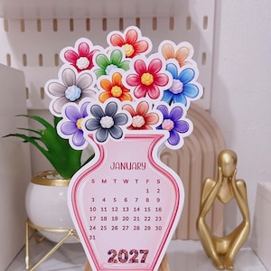 May include: A die-cut paper calendar for January 2027 shaped like a pink vase filled with colorful flowers. The vase features the month name, a grid of dates from 1 to 31, and the year 2027 at the bottom. The flowers are red, blue, orange, purple, and gray.