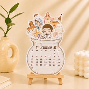 May include: A die-cut desk calendar for January 2027 featuring religious illustrations including Jesus, Mary, a praying child, a lamb, a cross, and a rosary. The calendar is shaped like a glass jar and rests on a small wooden easel stand.