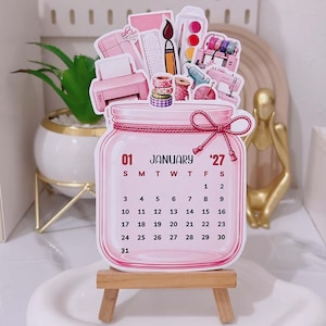 May include: A die-cut calendar for January 2027 shaped like a glass jar with a pink bow. The top features illustrations of craft supplies including a printer, ruler, paintbrush, watercolors, washi tape, and a sewing machine. It rests on a wooden easel.