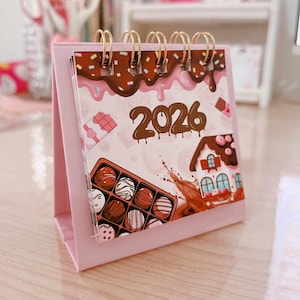 Puede incluir: Un calendario de escritorio rosa para el año 2026, con temática de chocolate. El calendario tiene encuadernación en espiral y un soporte rosa. Las páginas muestran ilustraciones de chocolate, incluyendo una casa de chocolate, una caja de bombones y goteo de chocolate.