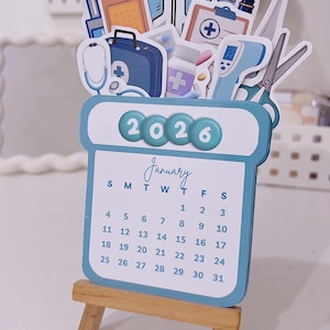 Puede incluir: Calendario azul y blanco de enero de 2026 con ilustraciones médicas, incluyendo un estetoscopio y un botiquín. El calendario se muestra en un pequeño caballete de madera.