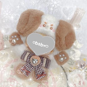 May include: A plush white and brown dog toy with a heart-shaped mirror that says "obabyo" on it. The toy is wearing a brown and white checkered bow with a rhinestone heart.