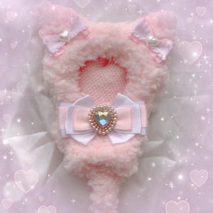 以下が含まれることがあります： パールと虹色のハート型の宝石が中央に付いた、大きく白とピンクのリボンが付いた、ピンクのふわふわした猫の形をしたぬいぐるみ。おもちゃの耳には、白いハートが付いたピンクのリボンが2つ付いています。