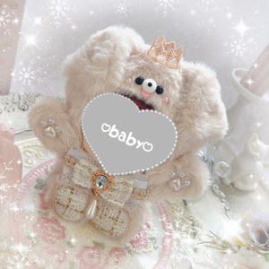 May include: A plush, light beige teddy bear with a gold crown, holding a gray heart-shaped sign with the text "obabyo". The bear has a decorative bow with a jewel and a pearl drop. The background has a soft, dreamy aesthetic with snowflake accents.