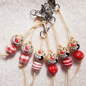 Könnte beinhalten: Eine Sammlung von Schlüsselanhängern mit clowngesichtigen Perlen und rot-weiß gemusterten Perlen. Jeder Schlüsselanhänger hat einen silberfarbenen Verschluss und ist an einer beigefarbenen Kordel aufgereiht. Die Clown-Gesichter haben rote Nasen und gemalte Smileys.