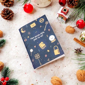 Peut inclure: Un carnet bleu foncé avec des illustrations dorées et le texte "Mon pense-bête pour le net" est entouré de décorations de Noël. Le carnet présente un cadenas, une clé et d'autres motifs fantaisistes. Noix, pommes de pin et baies rouges sont éparpillées.
