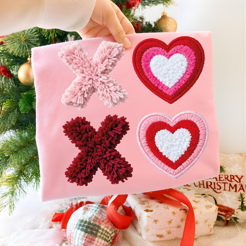 May include: A pink fabric square featuring a light pink and burgundy X and two embroidered hearts in red, pink, and white. The X's are made of fluffy yarn. The hearts have a layered design. The background includes a Christmas tree and wrapped gifts.