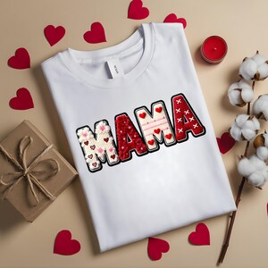 Op de afbeelding: Een wit t-shirt met het woord "MAMA" in een decoratief, gestikt ontwerp. De letters zijn rood en wit met hart- en bloemaccenten. Rode hartvormige uitsparingen en een kleine geschenkdoos zijn verspreid rond het shirt.