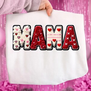 Op de afbeelding: Witte sweatshirt met het woord "MAMA" in een decoratief, gestructureerd ontwerp. De letters zijn zwart omlijnd en hebben verschillende patronen, waaronder harten, bloemen en een gebreide textuur in rood en wit. De sweatshirt wordt door een persoon omhoog gehouden.