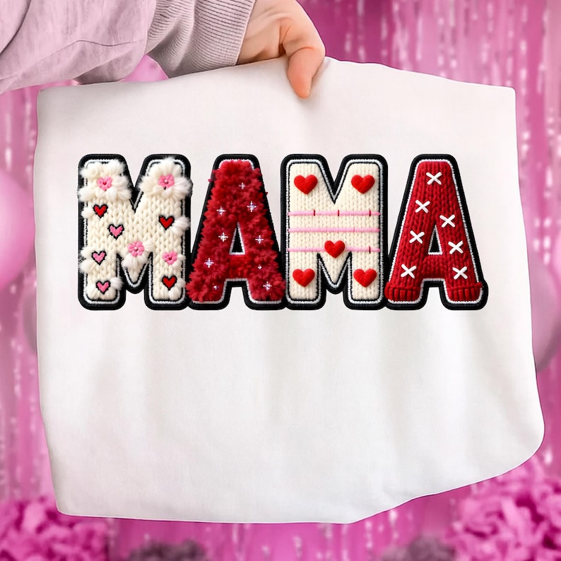 Op de afbeelding: Witte sweatshirt met het woord "MAMA" in een decoratief, gestructureerd ontwerp. De letters zijn zwart omlijnd en hebben verschillende patronen, waaronder harten, bloemen en een gebreide textuur in rood en wit. De sweatshirt wordt door een persoon omhoog gehouden.