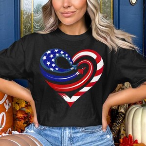 May include: Black t-shirt featuring a heart-shaped design with patriotic colors. The heart is composed of red and white stripes, blue with white stars, and a red airplane. The shirt is worn by a person.