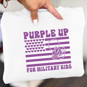 May include: White sweatshirt with a purple graphic. The graphic includes the text "PURPLE UP FOR MILITARY KIDS", a purple flag design with heart-shaped stars, and dog tags.