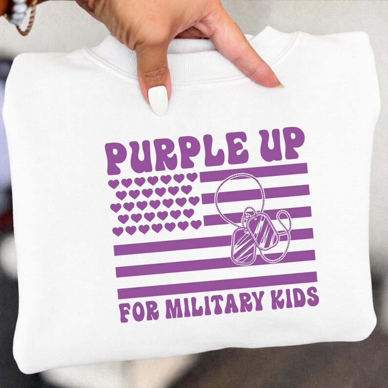 May include: White sweatshirt with a purple graphic. The graphic includes the text "PURPLE UP FOR MILITARY KIDS", a purple flag design with heart-shaped stars, and dog tags.