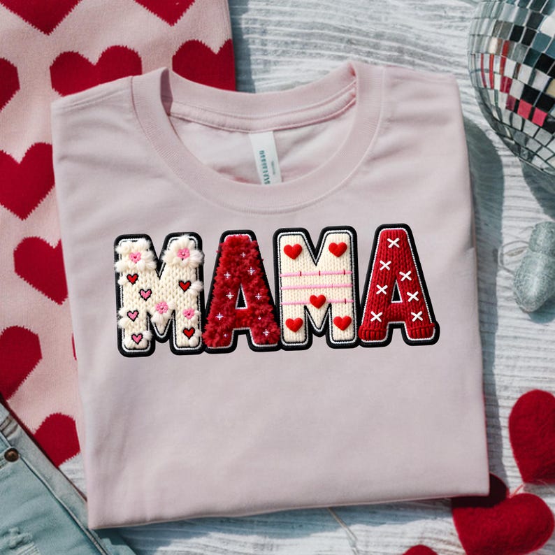 Op de afbeelding: Een lichtroze t-shirt met het woord "MAMA" in een decoratieve, geborduurde stijl. De letters zijn wit, rood en roze, met hart- en bloemaccenten. Het shirt is op een lichtgrijze ondergrond gevouwen.