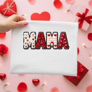 Op de afbeelding: Een witte draagtas met het woord "MAMA" in een decoratief, gestikt ontwerp. De letters zijn rood, wit en roze, met hart- en bloemaccenten. De tas wordt vastgehouden door een hand met roze nagellak, op een roze achtergrond.