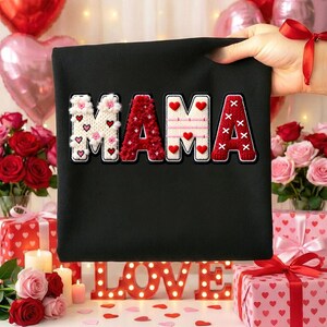 Op de afbeelding: Zwarte draagtas met het woord "MAMA" in een decoratief, gestikt ontwerp. De letters zijn wit, rood en cr&egrave;me, met hart- en X-accenten. De achtergrond bevat rozen, cadeaus, kaarsen en een lichtbord met de tekst "LOVE".