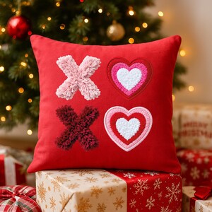 May include: A red decorative pillow with embroidered X's and O's. The X's are made of fluffy pink and burgundy yarn. Two hearts are embroidered with pink, red, and white accents. The pillow is placed on a gift box.