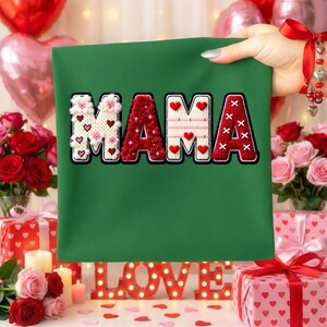 Op de afbeelding: Een groene stof met het woord "MAMA" geborduurd in wit, rood en roze. De letters zijn versierd met harten en bloemmotieven. De stof wordt door een hand omhoog gehouden, met op de achtergrond rozen, cadeaus en een "LOVE"-bord.
