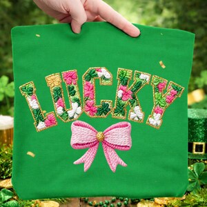 May include: Green tote bag with the word "LUCKY" embroidered in gold, pink, and white floral patterns. A pink bow is embroidered below the word. The bag is held up by a hand.
